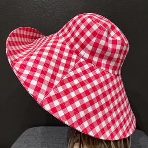 K&ouml;nnte beinhalten: Ein rot-wei&szlig; karierter Sonnenhut mit breiter Krempe. Der Hut ist aus Stoff gefertigt und hat ein klassisches Karomuster. Der Hut ist zum Schutz vor der Sonne gedacht.