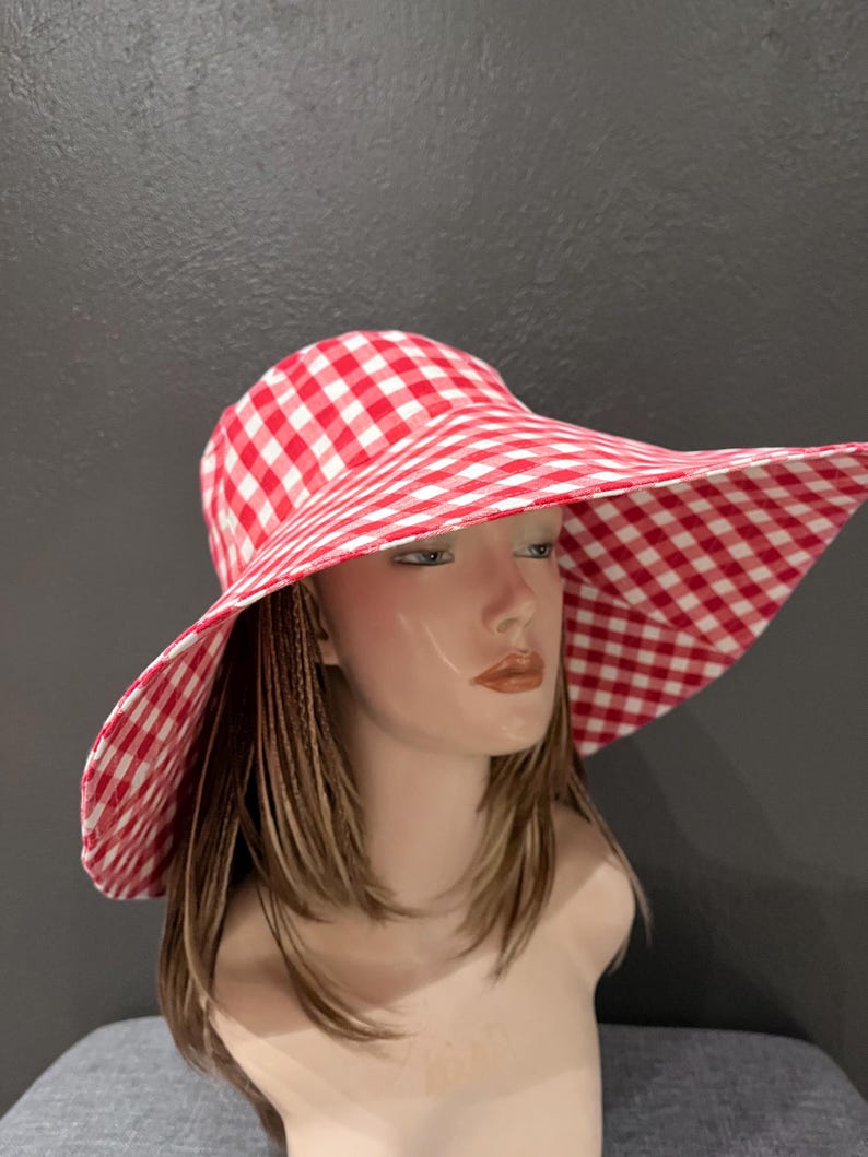 K&ouml;nnte beinhalten: Ein rot-wei&szlig; karierter Sonnenhut mit breiter Krempe. Der Hut wird auf einem Schaufensterkopf pr&auml;sentiert. Das Muster des Hutes ist ein klassisches Karomuster im Picknick-Stil.