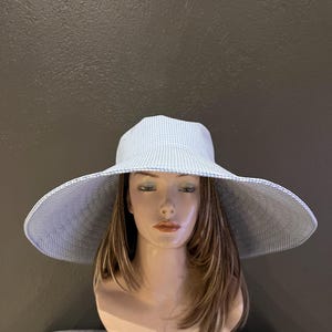 May include: A wide-brimmed sun hat with a light blue and white gingham pattern. The hat has a structured crown and a broad, floppy brim. The hat is displayed on a mannequin head with long, light brown hair.