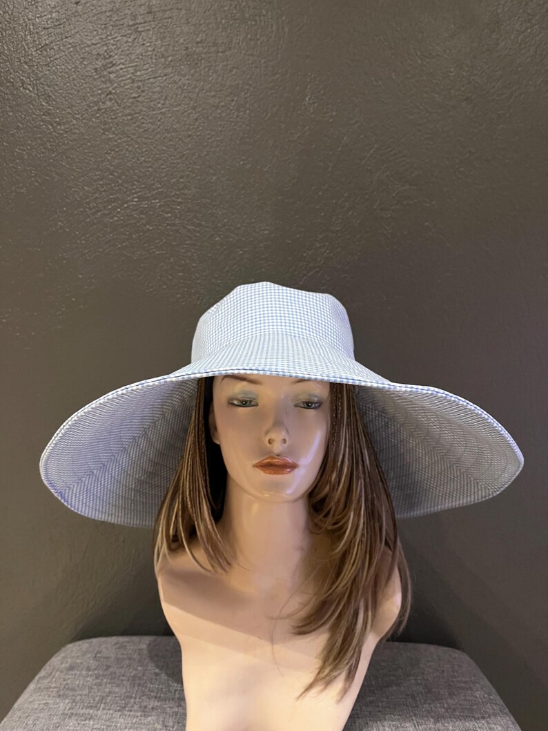 May include: A wide-brimmed sun hat with a light blue and white gingham pattern. The hat has a structured crown and a broad, floppy brim. The hat is displayed on a mannequin head with long, light brown hair.