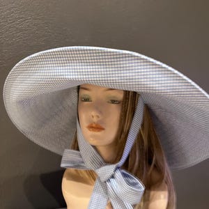 May include: A light blue and white gingham sun hat with a wide brim and a matching ribbon tie. The hat is displayed on a mannequin head. The ribbon is tied in a bow under the chin.