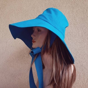 May include: A wide-brimmed sun hat in a bright turquoise blue. The hat has a large brim that extends out to the sides and a chin strap that ties under the chin. The hat is made of a lightweight fabric and is perfect for protecting your face and neck from the sun.