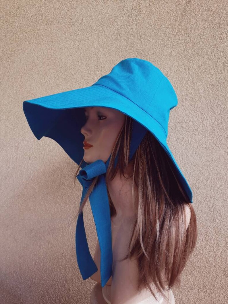 May include: A wide-brimmed sun hat in a bright turquoise blue. The hat has a large brim that extends out to the sides and a chin strap that ties under the chin. The hat is made of a lightweight fabric and is perfect for protecting your face and neck from the sun.