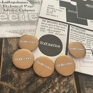 May include: Six gold-colored round buttons with the text "the Breeders" in white lettering. The buttons are arranged in a semi-circle on a wooden surface.