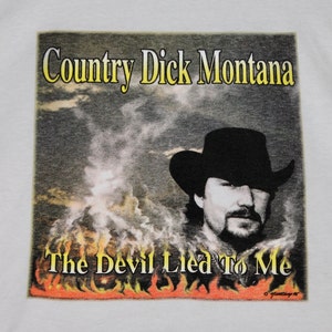 May include: A white t-shirt with a graphic print of a country music singer wearing a cowboy hat. The graphic is set against a background of clouds and flames. The text "Country Dick Montana" and "The Devil Lied To Me" are printed on the graphic.