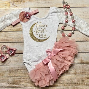May include: A white lace bodysuit with long sleeves and a gold glitter crescent moon with the text "Inara's first Eid". A pink tulle tutu with a pink satin bow. A pink flower headband and a pair of pink flower sandals.