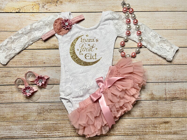 May include: A white lace bodysuit with long sleeves and a gold glitter crescent moon with the text "Inara's first Eid". A pink tulle tutu with a pink satin bow. A pink flower headband and a pair of pink flower sandals.