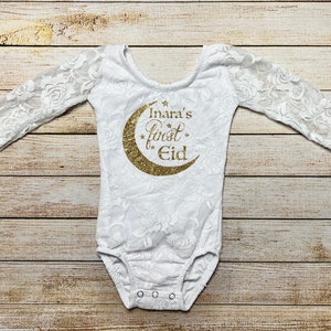 May include: White lace bodysuit with long sleeves and a gold glitter crescent moon design that says "Inara's First Eid".