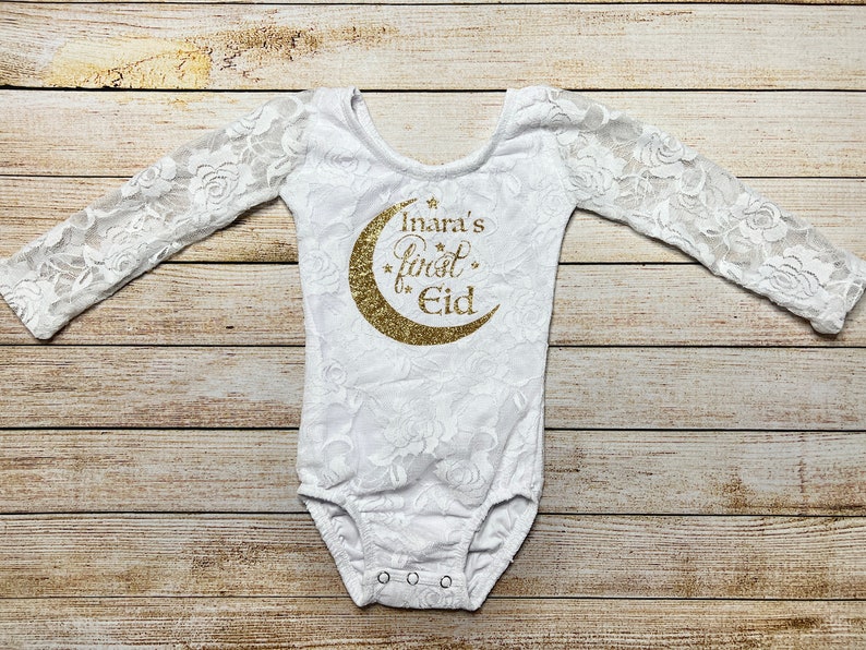 May include: White lace bodysuit with long sleeves and a gold glitter crescent moon design that says "Inara's First Eid".