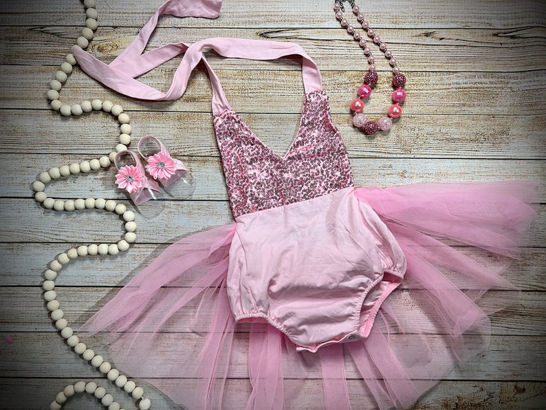 Puede incluir: Un conjunto de beb&eacute; ni&ntilde;a rosa con un corpi&ntilde;o de lentejuelas, falda de tul y zapatos a juego. El conjunto est&aacute; decorado con flores rosas.