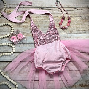Puede incluir: Un conjunto de beb&eacute; ni&ntilde;a rosa con un corpi&ntilde;o de lentejuelas, falda de tul y zapatos rosas a juego. El conjunto est&aacute; colocado sobre una superficie de madera con una guirnalda de cuentas blancas y un collar de cuentas rosas.