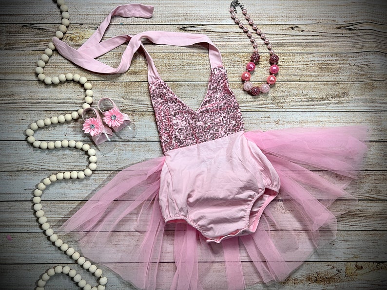 Puede incluir: Un conjunto de beb&eacute; ni&ntilde;a rosa con un corpi&ntilde;o de lentejuelas, falda de tul y zapatos rosas a juego. El conjunto est&aacute; colocado sobre una superficie de madera con una guirnalda de cuentas blancas y un collar de cuentas rosas.