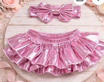 May include: A matching set of baby clothing in a shiny, metallic pink. The set includes a ruffled skirt and a headband with a large bow. The skirt has two layers of ruffles and an elastic waistband. The headband has a large bow.