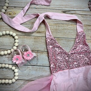 Puede incluir: Un vestido rosa con lentejuelas, falda de tul y top halter de sat&eacute;n rosa. El vestido se combina con zapatos de beb&eacute; de color rosa en forma de flor y un collar de cuentas rosas.