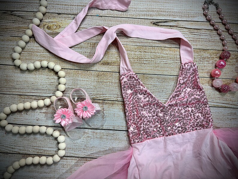 Puede incluir: Un vestido rosa con lentejuelas, falda de tul y top halter de sat&eacute;n rosa. El vestido se combina con zapatos de beb&eacute; de color rosa en forma de flor y un collar de cuentas rosas.