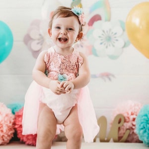 Puede incluir: Una ni&ntilde;a peque&ntilde;a con un disfraz de unicornio rosa y blanco con una diadema de flores. Est&aacute; de pie sobre un suelo de madera con un pomp&oacute;n rosa y azul en el fondo.