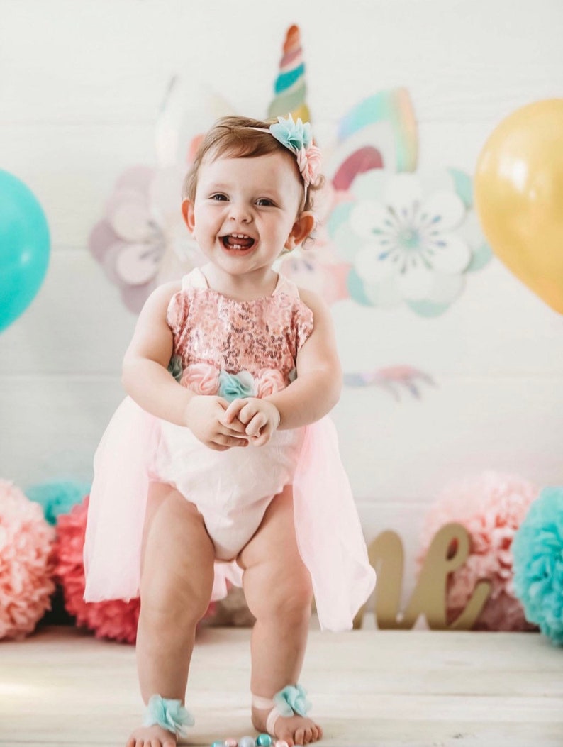 Puede incluir: Una ni&ntilde;a peque&ntilde;a con un disfraz de unicornio rosa y blanco con una diadema de flores. Est&aacute; de pie sobre un suelo de madera con un pomp&oacute;n rosa y azul en el fondo.