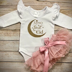 May include: White long-sleeved baby onesie with gold glitter crescent moon and stars design. The onesie says "Zayna's first Eid". A pink tulle tutu with a pink satin bow is included.
