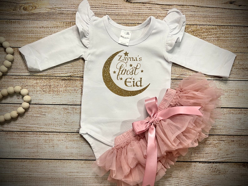 May include: White long-sleeved baby onesie with gold glitter crescent moon and stars design. The onesie says "Zayna's first Eid". A pink tulle tutu with a pink satin bow is included.
