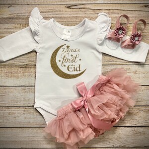May include: A white long-sleeved onesie with gold glitter lettering that reads "Zayna's First Eid" with a gold glitter crescent moon. The onesie is paired with a pink tulle skirt with a pink satin bow and a pair of pink sandals with a flower embellishment.