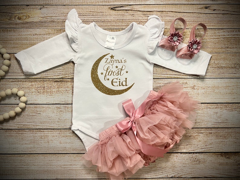May include: A white long-sleeved onesie with gold glitter lettering that reads "Zayna's First Eid" with a gold glitter crescent moon. The onesie is paired with a pink tulle skirt with a pink satin bow and a pair of pink sandals with a flower embellishment.