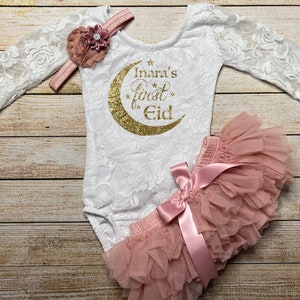 May include: A white lace bodysuit with long sleeves and a gold glitter crescent moon design. The text "Inara's First Eid" is printed on the bodysuit. A pink tulle tutu with a pink satin bow is included. A pink flower headband is also included.