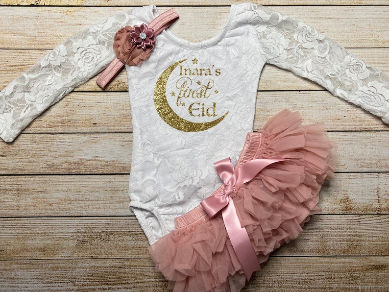 May include: A white lace bodysuit with long sleeves and a gold glitter crescent moon design. The text "Inara's First Eid" is printed on the bodysuit. A pink tulle tutu with a pink satin bow is included. A pink flower headband is also included.