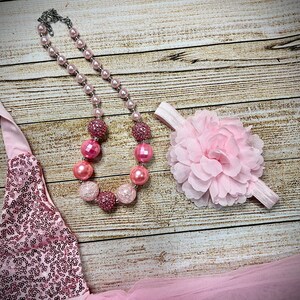 Puede incluir: Un vestido rosa de lentejuelas con escote halter, un collar de cuentas rosas con cuentas grandes y brillantes y una diadema de flores rosa.