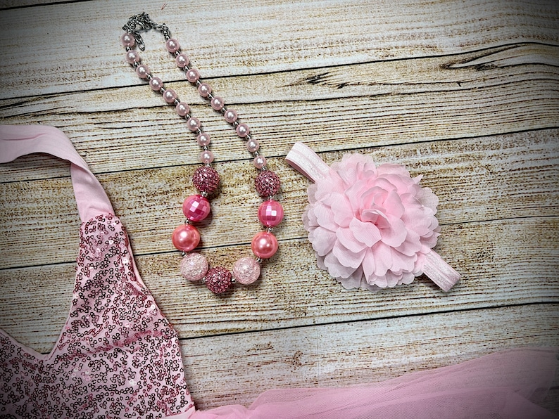 Puede incluir: Un vestido rosa de lentejuelas con escote halter, un collar de cuentas rosas con cuentas grandes y brillantes y una diadema de flores rosa.