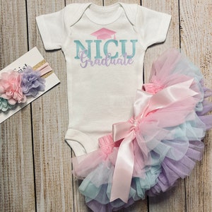 Puede incluir: Un body blanco con el texto "NICU Graduate" en letras azules y rosas con un gorro de graduación rosa sobre el texto. Una diadema de flores rosa, azul y morada y un tutú de tul rosa y azul con un lazo de satén rosa.