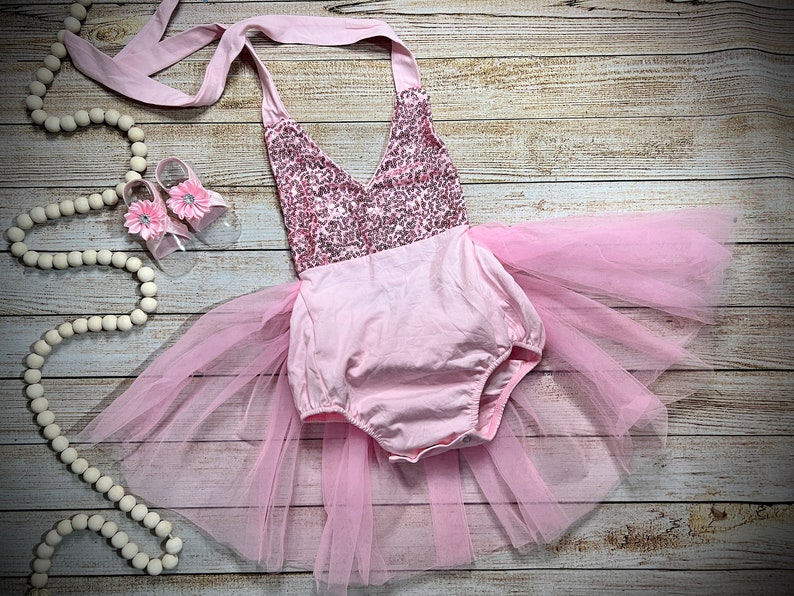 Puede incluir: Un conjunto de beb&eacute; ni&ntilde;a rosa con un corpi&ntilde;o de lentejuelas y una falda de tul. El conjunto est&aacute; colocado sobre una superficie de madera con un par de zapatos rosas y una guirnalda de cuentas blancas.