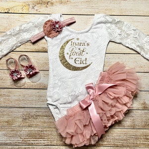 May include: A white lace bodysuit with long sleeves and a gold glitter crescent moon with the text "Inara's first Eid" on the front. The bodysuit is paired with a pink tulle skirt with a pink satin bow and a pair of pink sandals with flower embellishments.
