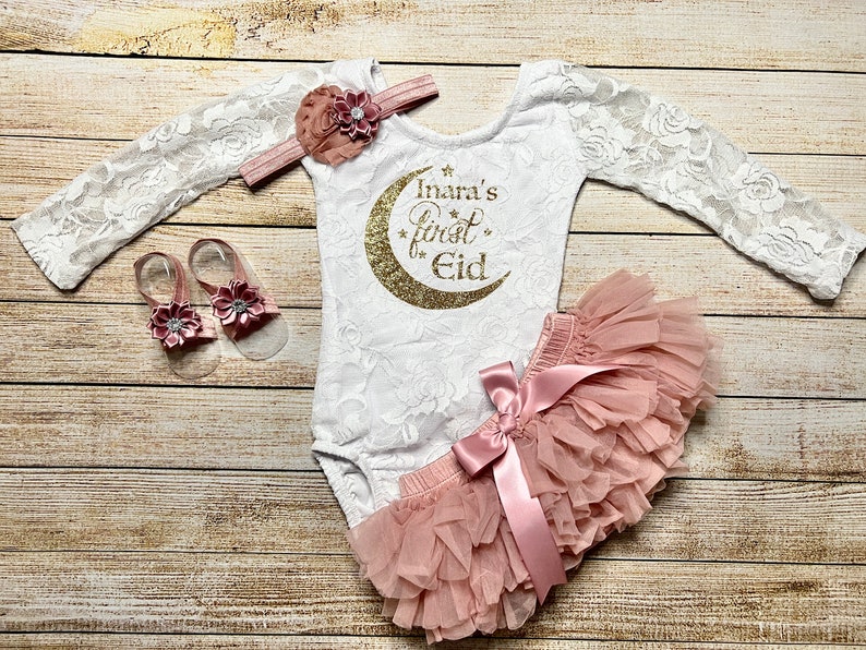 May include: A white lace bodysuit with long sleeves and a gold glitter crescent moon with the text "Inara's first Eid" on the front. The bodysuit is paired with a pink tulle skirt with a pink satin bow and a pair of pink sandals with flower embellishments.