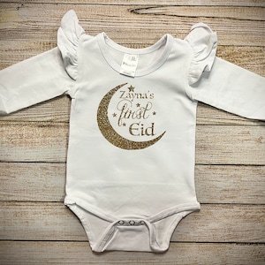 May include: White long-sleeved baby onesie with gold glitter crescent moon and stars graphic. The text "Zayna's first Eid" is written on the onesie.