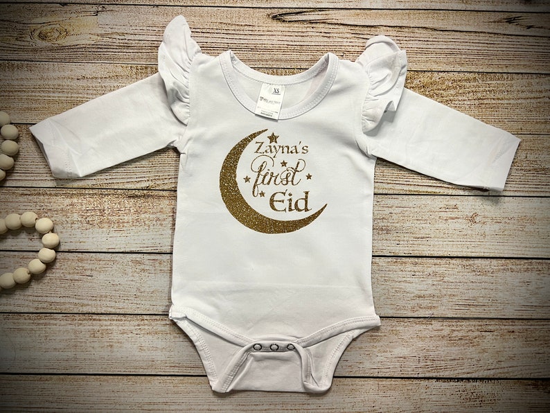 May include: White long-sleeved baby onesie with gold glitter crescent moon and stars graphic. The text "Zayna's first Eid" is written on the onesie.