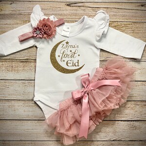 May include: A white baby onesie with gold glitter lettering that says "Zayna's first Eid" with a gold glitter crescent moon. The onesie has ruffled sleeves and is paired with a pink tulle skirt with a pink satin bow. A pink headband with a flower embellishment is also included.