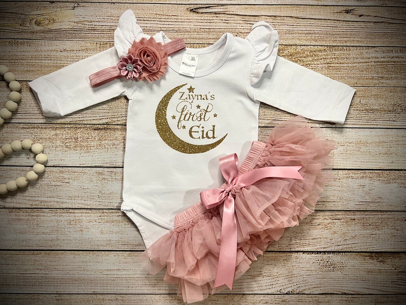 May include: A white baby onesie with gold glitter lettering that says "Zayna's first Eid" with a gold glitter crescent moon. The onesie has ruffled sleeves and is paired with a pink tulle skirt with a pink satin bow. A pink headband with a flower embellishment is also included.