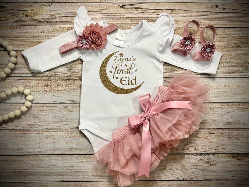 May include: A baby girl's outfit for Eid, featuring a white long-sleeved bodysuit with a gold glitter crescent moon and stars design that says "Zayna's First Eid", a pink tulle skirt with a pink satin bow, and a pair of pink sandals with flower embellishments.
