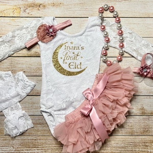 May include: A white lace bodysuit with a gold glitter crescent moon and the text "Inara's First Eid". The bodysuit is paired with a blush pink tulle skirt, a white lace headband with a blush pink flower, a blush pink pearl necklace, and blush pink flower sandals.