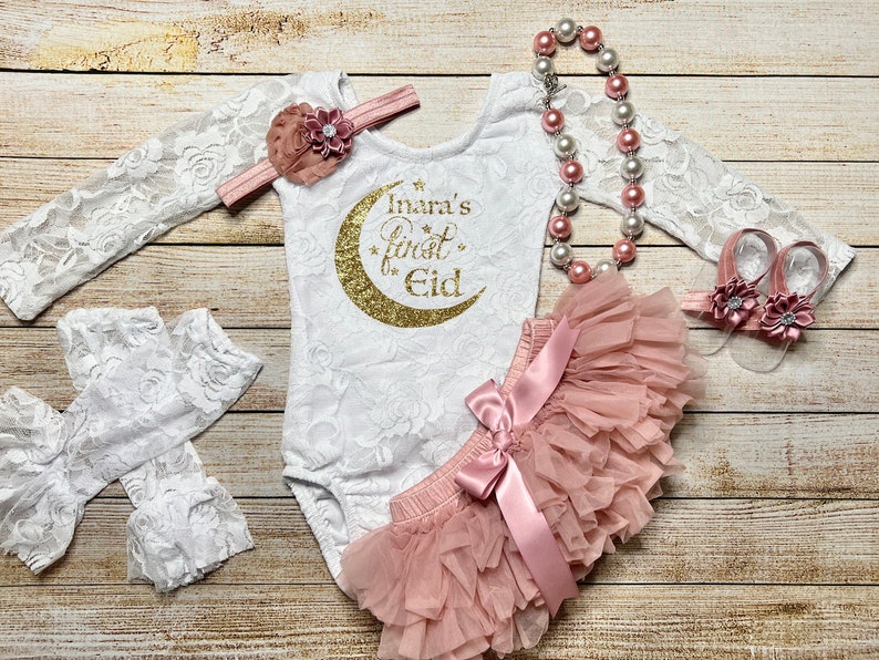 May include: A white lace bodysuit with a gold glitter crescent moon and the text "Inara's First Eid". The bodysuit is paired with a blush pink tulle skirt, a white lace headband with a blush pink flower, a blush pink pearl necklace, and blush pink flower sandals.
