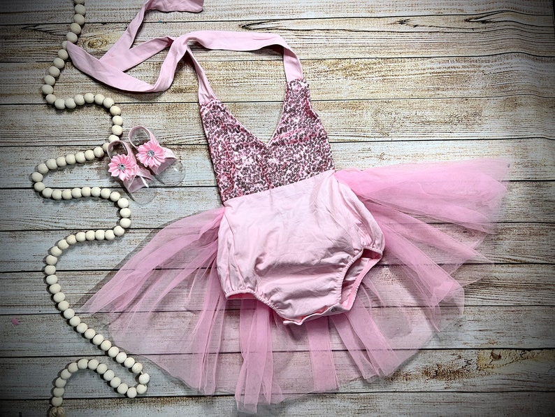 Puede incluir: Un conjunto de beb&eacute; ni&ntilde;a rosa con un corpi&ntilde;o de lentejuelas y una falda de tul. El conjunto est&aacute; colocado sobre una superficie de madera con una guirnalda de cuentas blancas y un par de sandalias rosas.