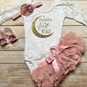 May include: A white lace bodysuit with long sleeves and a gold glitter crescent moon with the text "Inara's first Eid" on the front. The outfit is paired with a blush pink tulle tutu skirt with a satin bow and a matching headband with a blush pink flower. The set also includes a pair of blush pink sandals with a flower embellishment.