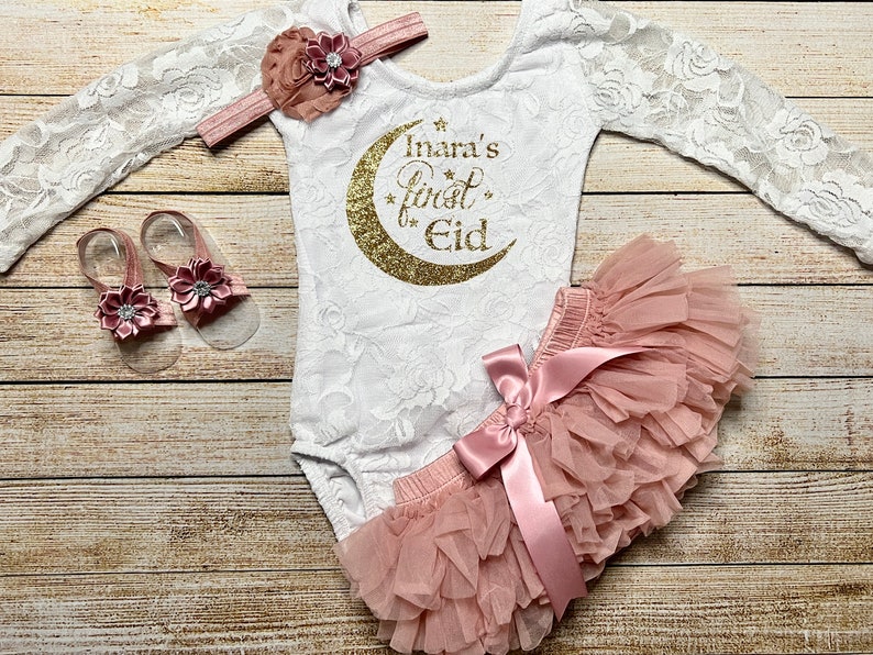 May include: A white lace bodysuit with long sleeves and a gold glitter crescent moon with the text "Inara's first Eid" on the front. The outfit is paired with a blush pink tulle tutu skirt with a satin bow and a matching headband with a blush pink flower. The set also includes a pair of blush pink sandals with a flower embellishment.