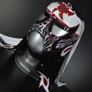Puede incluir: Una m&aacute;scara de lucha libre con un dise&ntilde;o llamativo. La m&aacute;scara presenta una base blanca con detalles rojos y negros, incluyendo un estampado de cebra en los laterales. La m&aacute;scara est&aacute; adornada con lentejuelas y tiene una superficie brillante y reflectante.