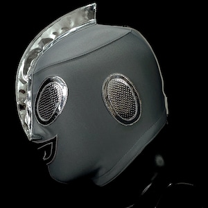 May include: A gray wrestling mask with silver accents. The mask features large, oval eye openings with a textured silver finish and a silver trim. A silver trim also outlines the top of the mask. The mask has a smiling mouth design.