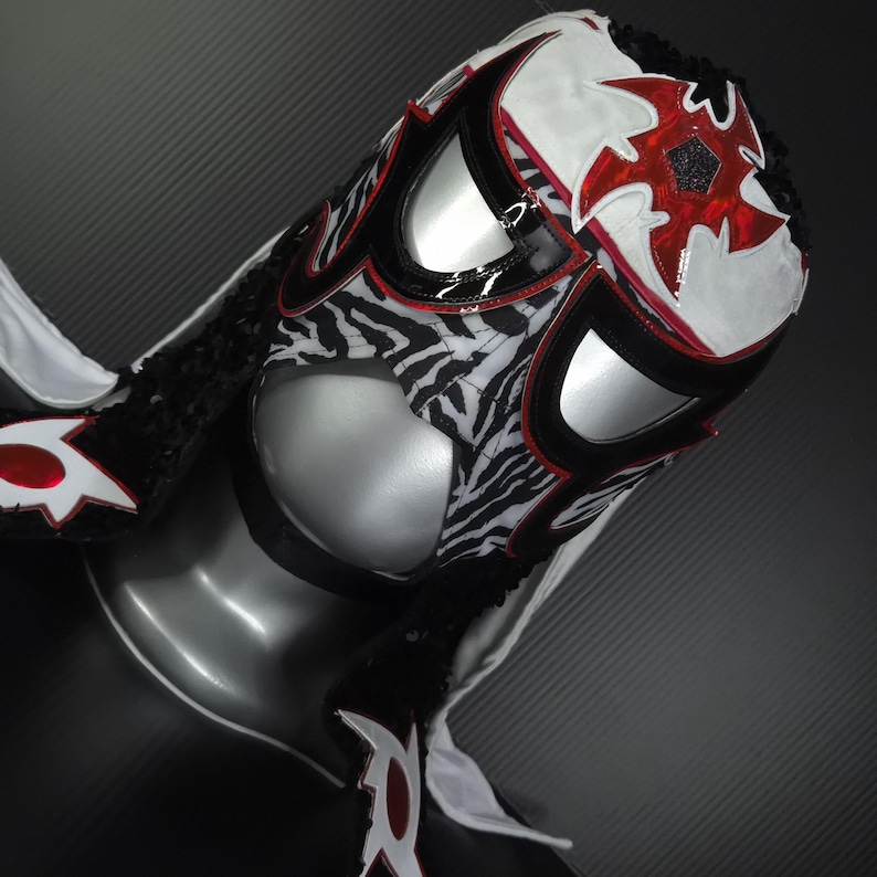 Puede incluir: Una m&aacute;scara de lucha libre con un dise&ntilde;o llamativo. La m&aacute;scara presenta un patr&oacute;n de cebra en la parte inferior de la cara, con detalles en blanco y rojo. La parte superior es blanca con formas rojas en forma de estrella. Ribete negro y cubiertas oculares plateadas.