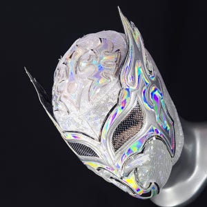 Op de afbeelding: Een zilveren lucha libre-masker met een holografisch, iriserend ontwerp. Het masker heeft vlamachtige accenten en ingewikkelde details, met een mesh-stof voor het gezicht. Het masker wordt getoond op een mannequinhoofd tegen een zwarte achtergrond.