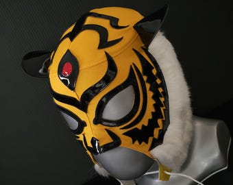 May include: A yellow and black wrestling mask with a tiger design. The mask features black accents, a red eye detail, and a white fur trim around the neck. The mask is displayed on a mannequin head.