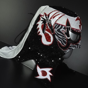 Puede incluir: Una m&aacute;scara de lucha libre profesional con un dise&ntilde;o blanco, rojo, negro y plateado. La m&aacute;scara presenta un drag&oacute;n y formas geom&eacute;tricas, con un patr&oacute;n de cebra en el lateral. La m&aacute;scara est&aacute; cubierta de lentejuelas negras y tiene una capa blanca.