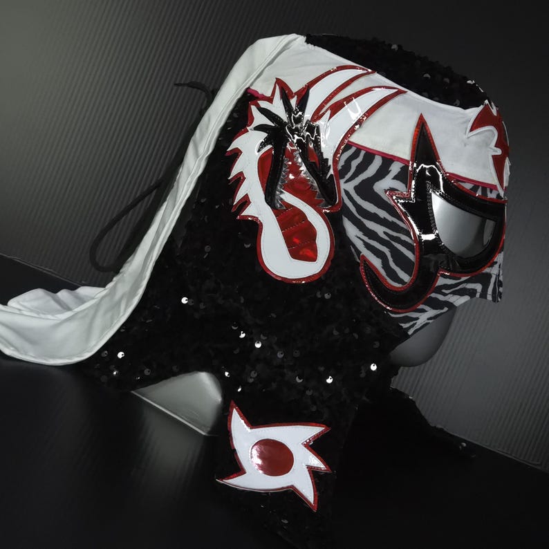 Puede incluir: Una m&aacute;scara de lucha libre profesional con un dise&ntilde;o blanco, rojo, negro y plateado. La m&aacute;scara presenta un drag&oacute;n y formas geom&eacute;tricas, con un patr&oacute;n de cebra en el lateral. La m&aacute;scara est&aacute; cubierta de lentejuelas negras y tiene una capa blanca.